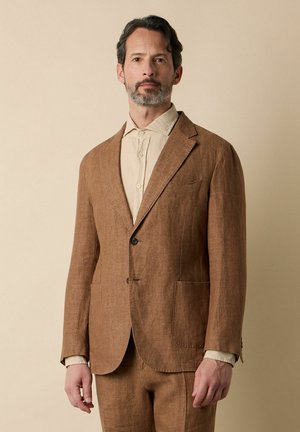 Mann mit kurzen dunklen Haaren und Bart, trägt ein hellbeiges Hemd und braunen Leinenanzug, stehend vor einem einfarbigen beigen Hintergrund.