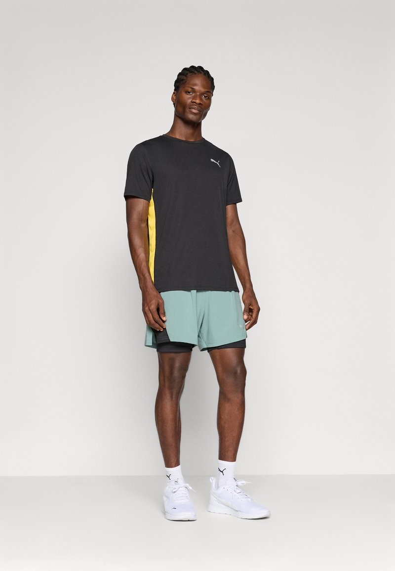 Camiseta atlética negra con panel lateral amarillo, combinada con pantalones cortos verde menta. El modelo lleva zapatillas blancas y calcetas deportivas, de pie sobre un fondo gris.
