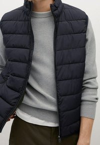 Granatowa pikowana kamizelka z wysokim kołnierzem, zamkiem błyskawicznym i bocznymi kieszeniami. Noszona na jasnoszarim prążkowanym swetrze. Gładka faktura i strukturalny krój.