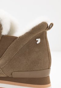 Chaussure en daim marron avec doublure en fausse fourrure, au design arrondi, et semelle en caoutchouc beige avec un léger talon et un accent de logo métallique.