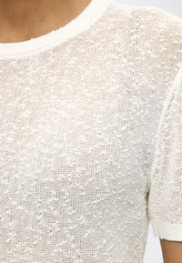 Κοντινό πλάνο ενός ατόμου που φοράει λευκό πλεκτό τοπ με υφή, κοντά μανίκια και στρογγυλή λαιμόκοψη.