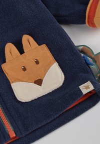 Giacca in pile blu navy con una tasca a forma di volpe marrone dotata di orecchie, zip arancione e fodera rossa. Tessuto testurizzato con un elemento di design giocoso.