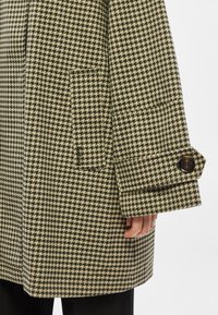 Manteau en laine à carreaux crème et vert foncé, avec une poche plaquée et des poignets boutonnés. Tissu texturé et silhouette structurée.