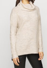 Beige gebreide trui met een col, lange mouwen en zijsplitten. Zachte textuur en losse pasvorm, met een geribbeld patroon over het geheel.