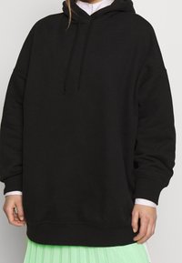 Felpa nera oversize realizzata in un materiale morbido, con cappuccio regolabile e polsini a coste. Indossata sopra una camicia bianca e una gonna plissettata verde menta.