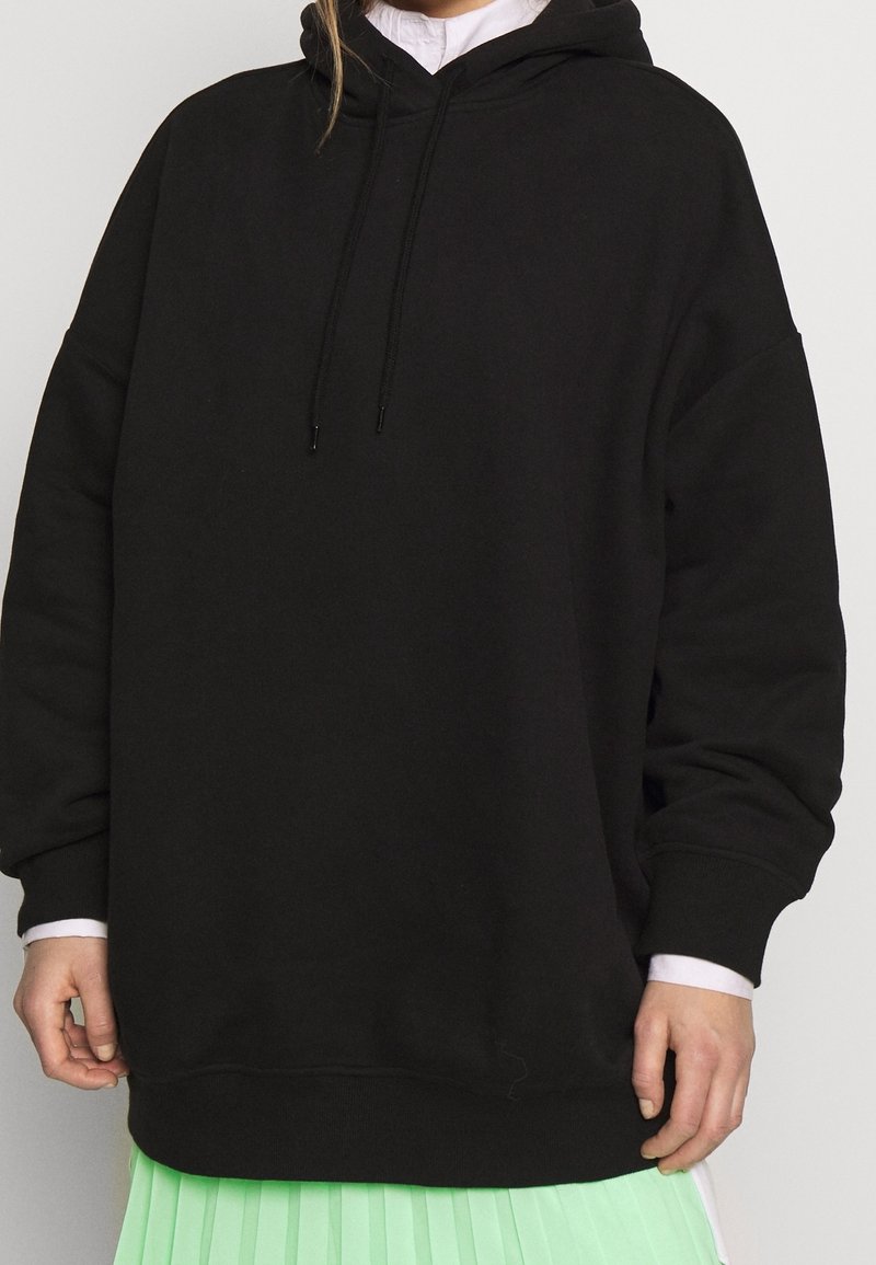 Felpa nera oversize realizzata in un materiale morbido, con cappuccio regolabile e polsini a coste. Indossata sopra una camicia bianca e una gonna plissettata verde menta.