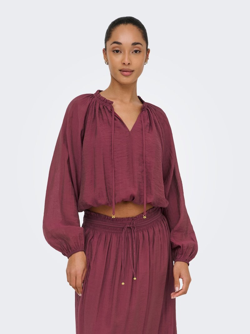 Γυναίκα που φοράει μακρυμάνικο βυσσινί crop top με κορδόνι στη λαιμόκοψη και ταιριαστή φούστα με ψηλή μέση σε απλό λευκό φόντο.