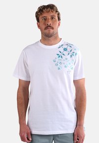 T-shirt en coton blanc avec un motif floral bleu sur l'épaule gauche. Coupe ample, col rond, manches courtes et texture lisse.