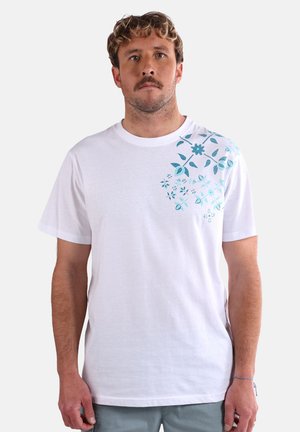 T-shirt en coton blanc avec un motif floral bleu sur l'épaule gauche. Coupe ample, col rond, manches courtes et texture lisse.