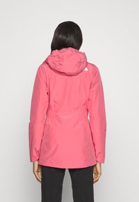Chaqueta impermeable rosa con capucha, cintura ajustable mediante cordón y puños elásticos. Presenta un logo en la parte superior del respaldo y una textura suave y ligera.