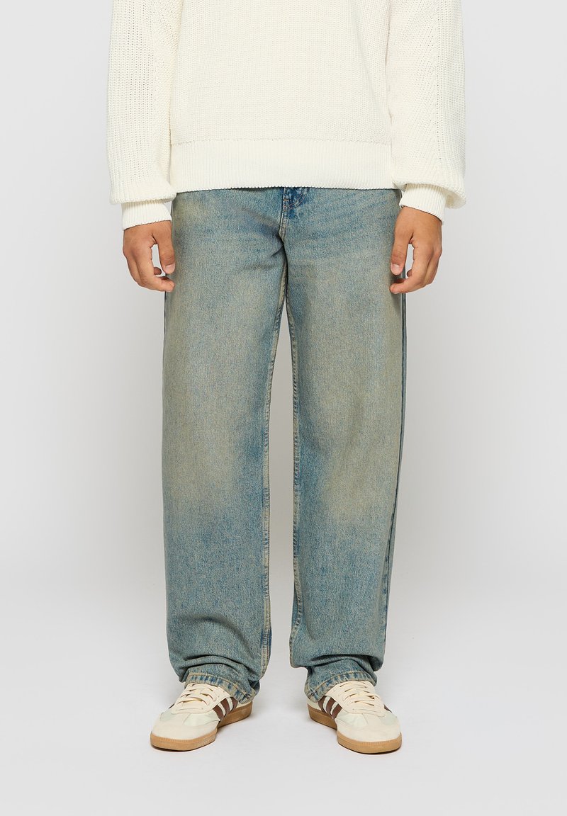 Lyseblå, vide bukser i denim med en klassisk denimstruktur, parret med en cremefarvet strikket sweater og beige sneakers med mørkebrune striber.