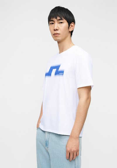 Νεαρός άνδρας που φοράει λευκό μπλουζάκι με μπλε σχέδιο και ανοιχτόχρωμα μπλε τζιν, στέκεται μπροστά σε απλό ανοιχτό φόντο.