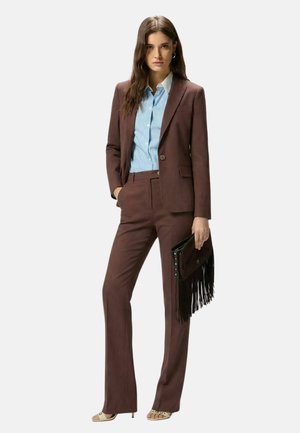 Donna con un completo marrone su misura e una camicia azzurro chiaro a righe, che tiene una pochette nera con frange, in piedi con una mano in tasca.