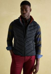 Mann in einer schwarzen gesteppte Weste über einem hochgekrempelten karierten Hemd und roten Hosen, der vor einem beigen Hintergrund steht.