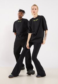 Doi modele poartă tricouri negre, supradimensionate, cu inscripția "New York" în verde neon. Modelul masculin le asortează cu pantaloni negri și încălțăminte masivă; modelul feminin poartă flare negre cu textură ribbed.