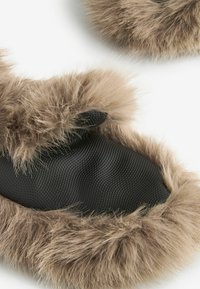 Faux fur -tossut, joissa on pehmeä, beige pörröinen pinta ja tekstiilinen musta materiaali sisällä. Pyöreä muoto, jossa on ylellinen reunus.