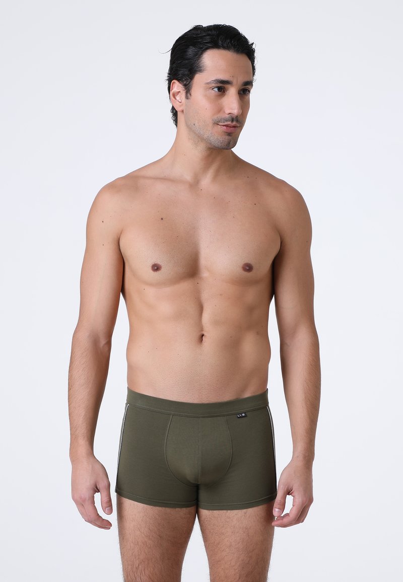 Uomo senza maglietta con capelli scuri, che indossa boxer verde oliva, in piedi contro uno sfondo grigio chiaro uniforme, rivolto leggermente verso destra.