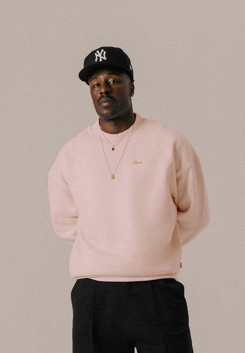 Sweat-shirt rose clair avec un col rond et des poignets côtelés. Présente un logo subtil en or et est associé à un pantalon noir et une casquette noire.
