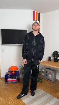 Fekete zipzáros kapucnis pulóver csillagmintával, fekete nadrággal párosítva. Fekete cipő fehér csíkokkal. A szobában TV és szobanövények találhatóak.