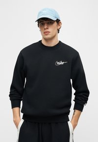 Νέος άνδρας που φοράει μαύρο φούτερ Nike και μαύρο παντελόνι με λευκές ρίγες, ανοικτό μπλε καπέλο Nike, στέκεται με τα χέρια στις τσέπες.