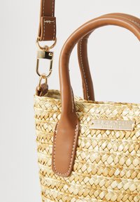 Sac en paille tissé avec des poignées en cuir marron et une bandoulière amovible. Présente une plaque de logo dorée et des accents brodés sur les poignées.