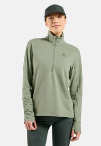 Helltürkises Quarter-Zip Langarmshirt aus strukturiertem Stoff mit Daumenlöchern, kombiniert mit passenden, figurbetonten Hosen und einer grünen Mütze.