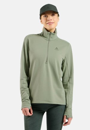 Helltürkises Quarter-Zip Langarmshirt aus strukturiertem Stoff mit Daumenlöchern, kombiniert mit passenden, figurbetonten Hosen und einer grünen Mütze.