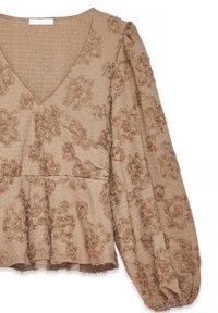 Blouse beige texturée avec col en V, manches longues bouffantes, motif floral en relief et bas à basque.