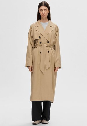 Beige trench coat s dvojitým zapínáním, černými knoflíky a páskem v pase, zkombinovaný s širokými černými kalhotami a béžovými balerínami.