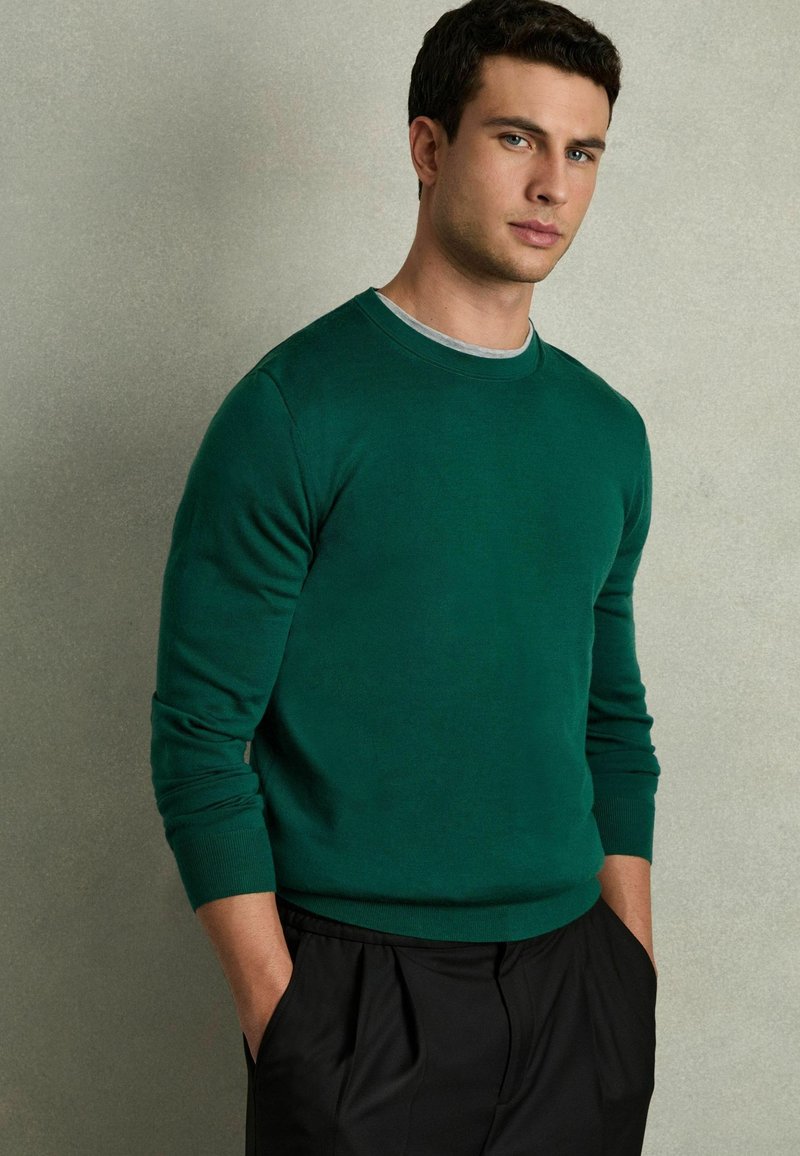 Zöld crewneck pulóver, puha anyagból, hosszú ujjakkal, texturált felülettel és bordás mandzsettákkal. Egy világosszürke ing felett viselve.