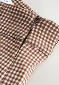 Primer plano de una tela a cuadros marrón y blanca con una cremallera negra y una lengüeta de tela en una prenda.