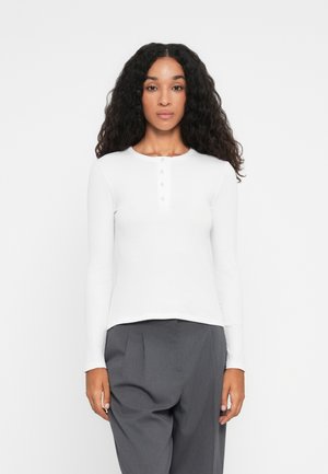 Hvit stripete langermet topp med rund hals og fem-knaps placket, kombinert med grå plissert bukser. Glatt tekstur og tettsittende design.