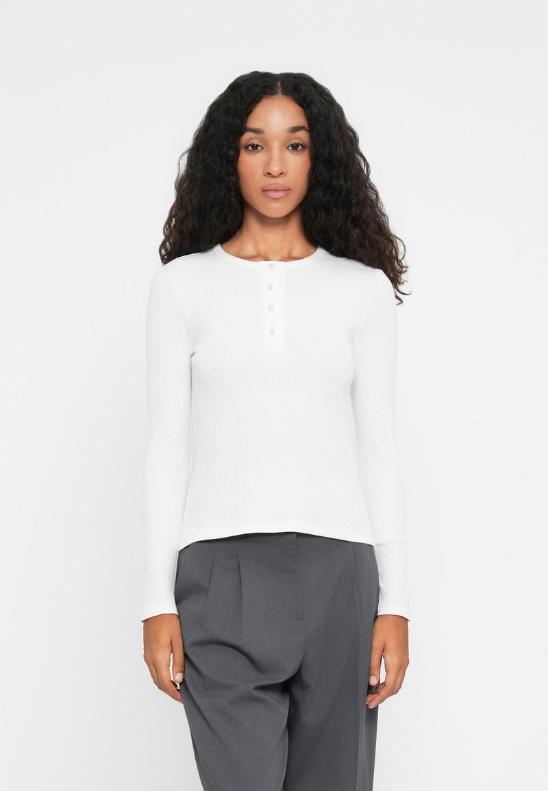 Haut long à manches blanches en côtes avec un col rond et une patte de boutonnage à cinq boutons, associé à un pantalon plissé gris. Texture lisse et design ajusté.