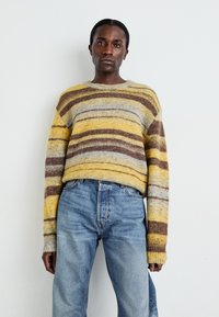 Suéter de rayas en amarillo, gris y marrón con mangas largas, combinado con jeans de mezclilla azul claro. Textura suave, corte relajado.