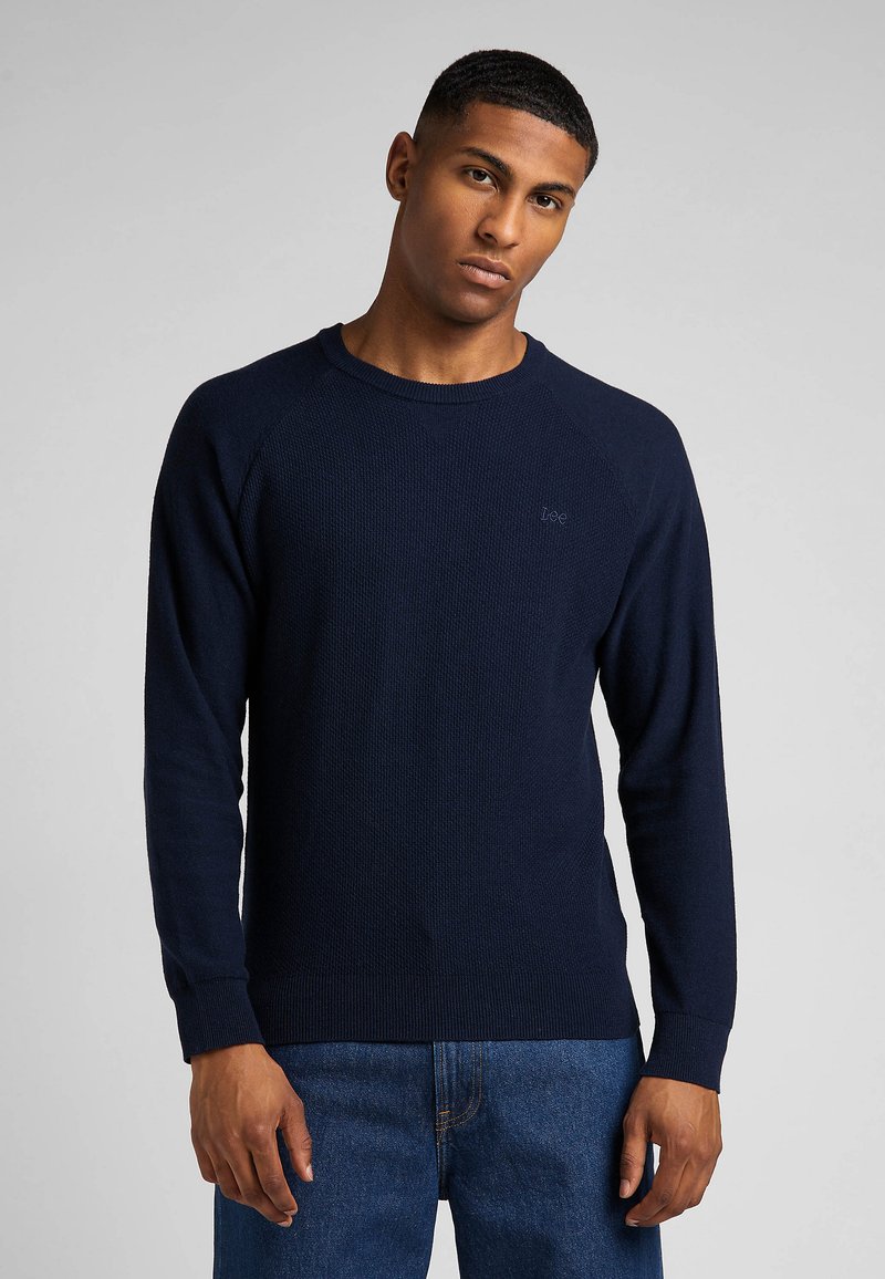 Pull en maille côtelée bleu marine à manches longues et col rond, avec un logo discret sur la poitrine et une coupe décontractée.