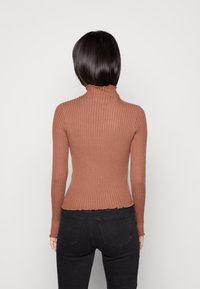 Maglione a dolcevita marrone a coste con maniche lunghe, presenta un colletto arricciato e una finitura testurizzata, abbinato a jeans neri aderenti.