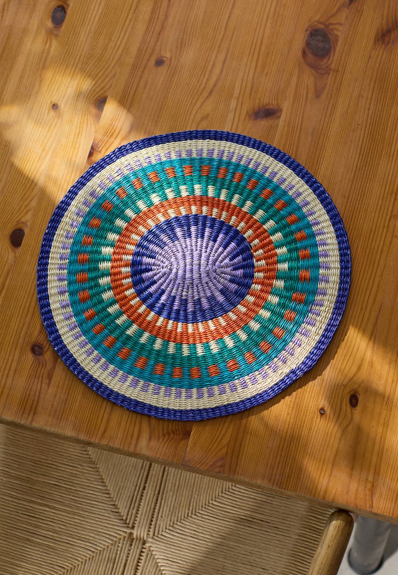Panier tressé rond avec un motif multicolore incluant des teintes vertes, orange, violettes et crème. Surface texturée avec des détails rayés.