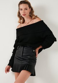 Suéter negro de canalé con escote tipo off-shoulder combinado con una minifalda de cuero negra de cintura alta, con dobladillo plisado y cierre de botones.
