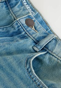 Lyseblå cowboybukser i denim med knaplukning, synlige syninger og en tilspidset pasform. Viser slidt tekstur og mærketag.