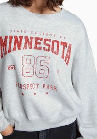 Vaaleanharmaa collegepaita, jossa on punainen graafinen teksti "STATE COLLEGE OF MINNESOTA 86 PROSPECT PARK." Paidassa on resorit hihansuissa ja helmassa, ja sen materiaali on pehmeää.
