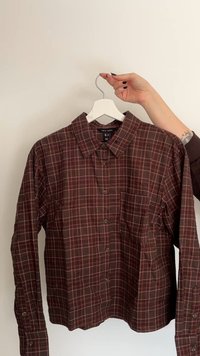 Camicia marrone a quadri con bottoni, maniche lunghe, colletto e taschino sul petto; presenta motivi lineari rossi e arancioni. Appesa a una gruccia bianca.