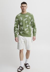 Groene sweater met bloemenprint, geribde boorden en zoom. Gecombineerd met lichtgrijze shorts, witte sokken en bruine sandalen. Minimalistisch ontwerp.