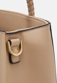 Beżowa torebka wykonana z ekologicznej skóry, z plecioną rączką, złotymi akcentami i czarnym wykończeniem wzdłuż górnej krawędzi. Gładka tekstura z żelazną formą.