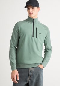 Minzgrüner Zip-Up-Pullover mit hohem Kragen und schwarzem Reißverschluss. Taillierte Bündchen und Saum mit Rippstrick. Mit subtiler Markenplatzierung auf der Brust.