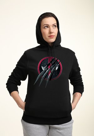 Kobieta w czarnej bluzie z kapturem z pazurami Wolverina na czerwonym symbolu Deadpool, ręce w kieszeniach, patrzy w górę w prawo.