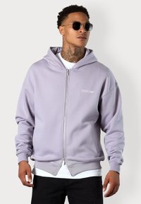 Light purple hooded sweatshirt with front zipper, ribbed cuffs and hem, featuring a white logo on the chest, layered over a white shirt.