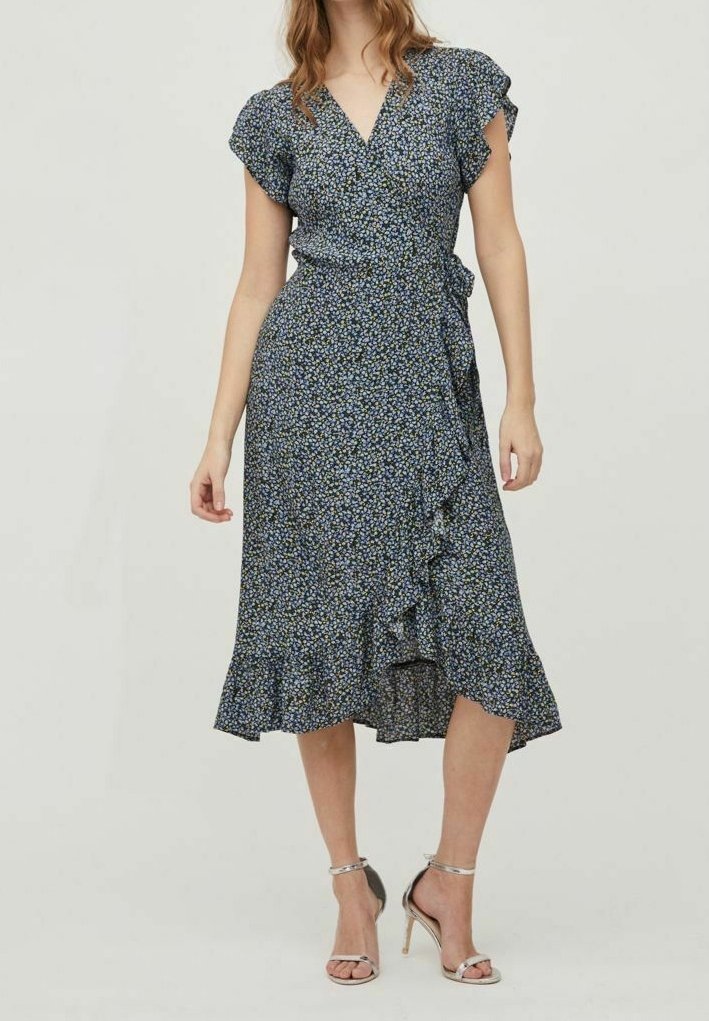 Femme portant une robe portefeuille fleurie bleu marine avec des bords volantés et des sandales argentées à talons hauts, debout sur un fond clair.