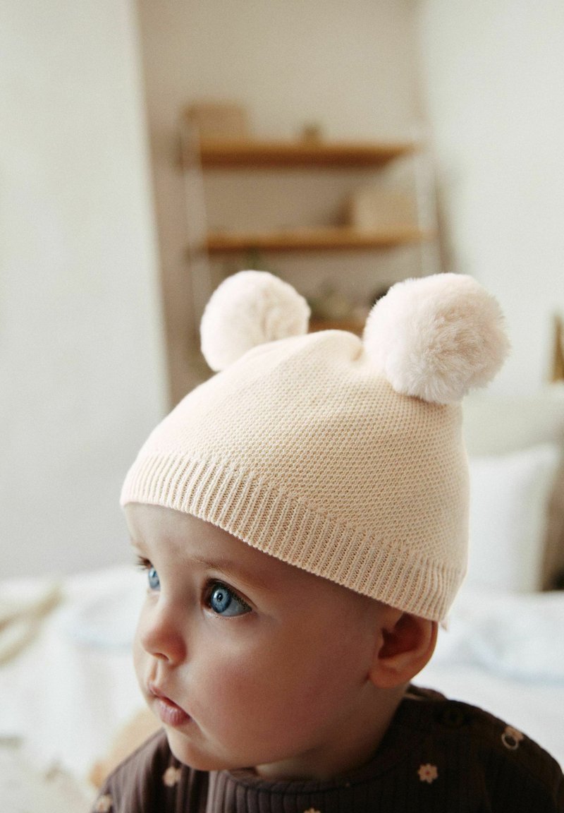 Cappello lavorato a maglia beige chiaro con due pompon soffici in cima. Presenta un orlo a coste e un motivo testurizzato.