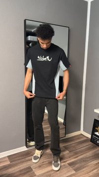 Joven ajustando una camiseta negra y gris con texto blanco, de pie frente a un espejo alto en una habitación con suelo de madera y paredes grises.