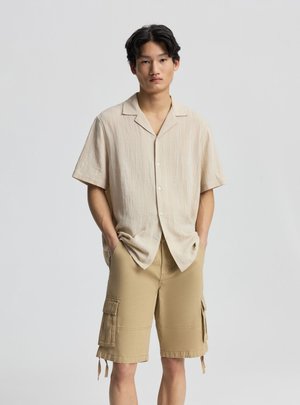 Giovane uomo con capelli neri che indossa una camicia beige a maniche corte con bottoni e pantaloncini cargo color sabbia, in piedi con le mani nelle tasche.